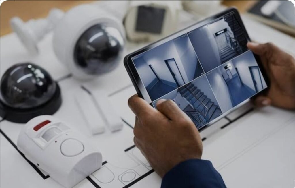 "A hands-on demonstration of a modern smart security and CCTV surveillance monitoring setup used to safeguard the Apex International School campus. A technician or safety officer holds a digital tablet displaying a multi-camera split-screen grid view showing clear real-time footage of indoor hallways, stairwells, and secure school corridors. Arrayed on the workspace table next to the tablet are various high-tech security components, including multiple dome and bullet CCTV cameras, motion sensors, and wireless security units resting on a blueprint layout. This image highlights the advanced student safety protocols, 24/7 campus surveillance, and secure environment maintained across the premier school and coaching facilities in the Manigachhi and Darbhanga region."