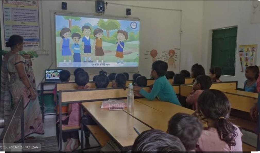 "An active learning session inside a smart digital classroom at Apex International School. A female teacher stands at the front of the room near a large projection screen displaying an interactive, colorful educational cartoon setup for young learners. Students are seated at wooden desks and benches, facing the audio-visual projection display. The room is equipped with electronic speakers, a laptop connection terminal, wall charts, and a green chalkboard. This image highlights the advanced infrastructure, digital classrooms, and audio-visual teaching aids used at the premier school and coaching facilities in the Manigachhi and Darbhanga region."