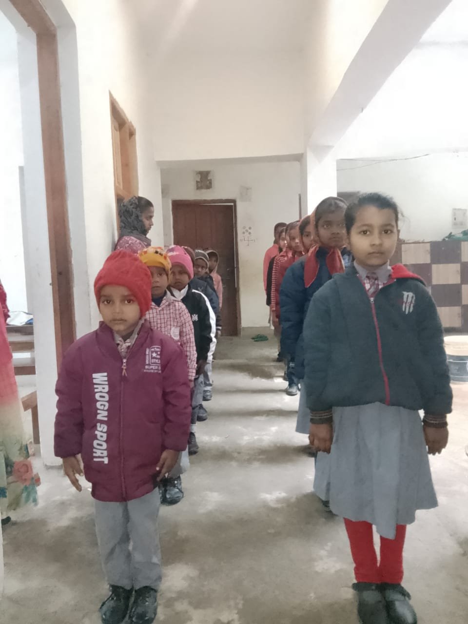 "A real-life campus photo showing students of Apex International School standing disciplined in structured lines along an indoor corridor. The children are dressed in winter school uniforms, wearing dark navy blue zip-up jackets, maroon hoodies, and beanies. The school crest is visible on the jackets of the senior students. The corridor features white walls, tiled accents, and polished flooring, reflecting a safe, clean, and organized environment. This image captures daily school discipline, student life, and the structural childhood care offered at the premier school and coaching facilities in the Manigachhi and Darbhanga region."